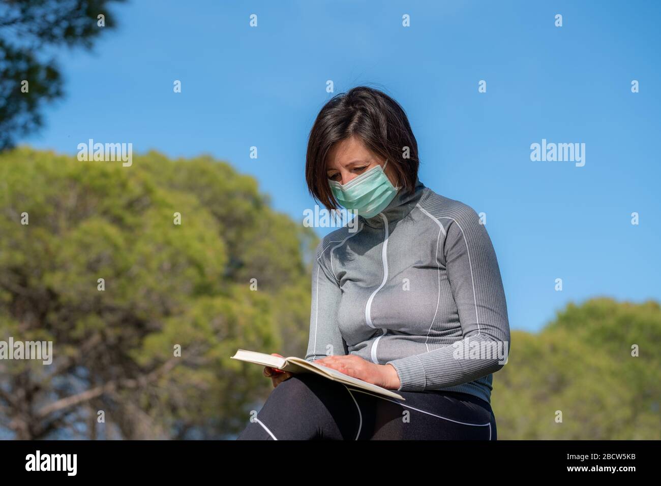 Ragazza nel periodo di un coronavirus in una maschera che legge un libro in natura. Isolamento sociale. Rimani a casa. Foto Stock