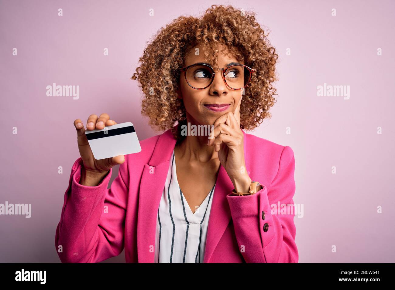 Giovane afro-americana business woman che tiene identificazione carta di identità su sfondo rosa serio faccia pensare alla domanda, molto confuso idea Foto Stock