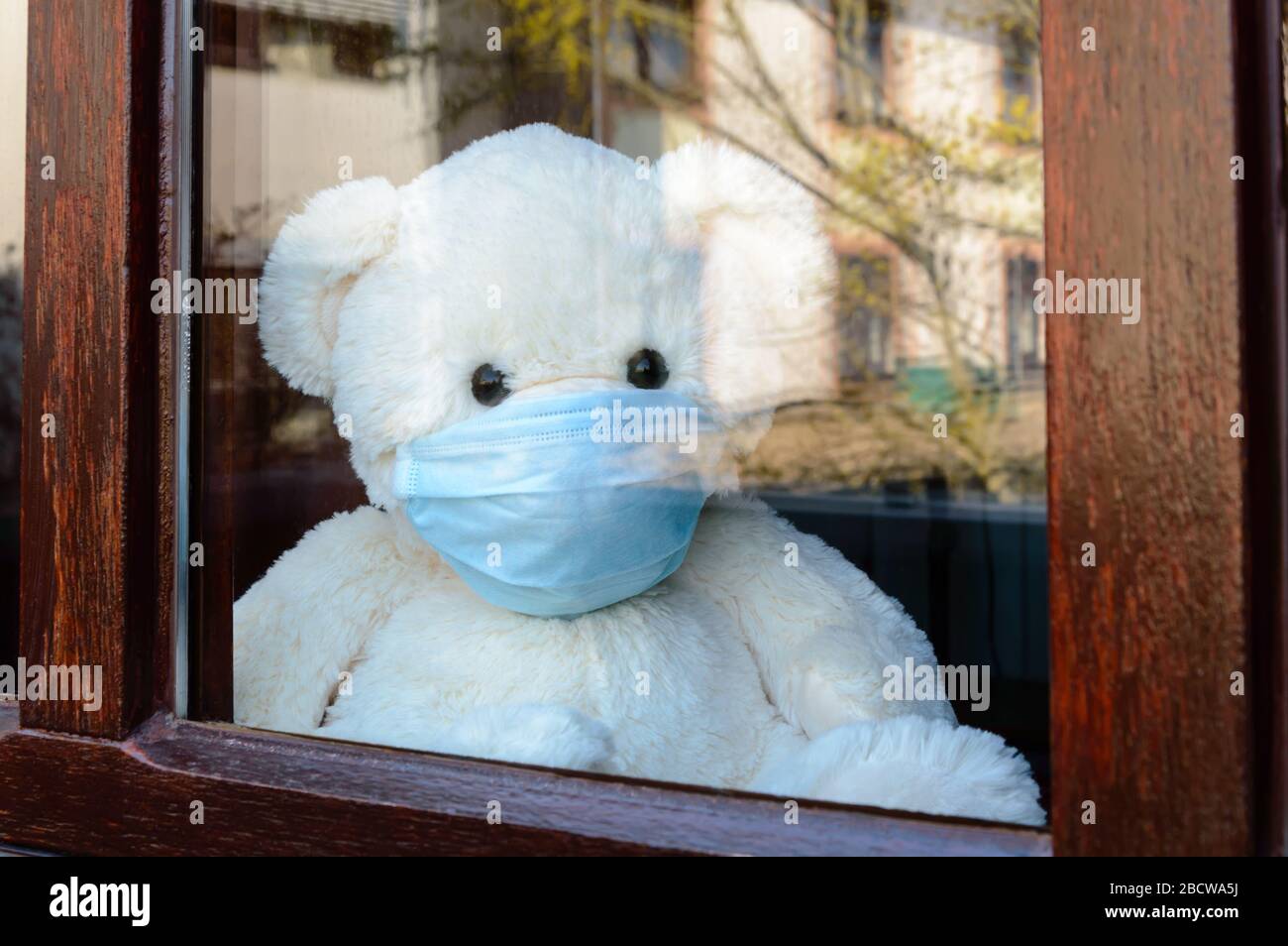 Simpatico orsacchiotto triste con maschera medica protettiva in quarantena per Covid-19 si affaccia dalla finestra. Concetto di soggiorno a casa. Malattia, isolamento del paziente e. Foto Stock
