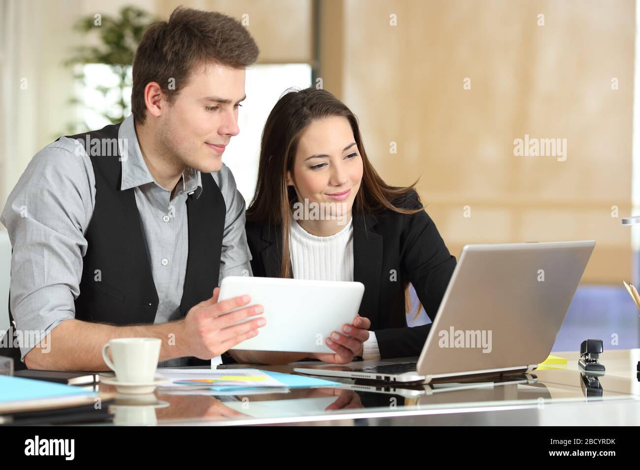 Due dipendenti confrontano i contenuti dei tablet e dei notebook in ufficio Foto Stock