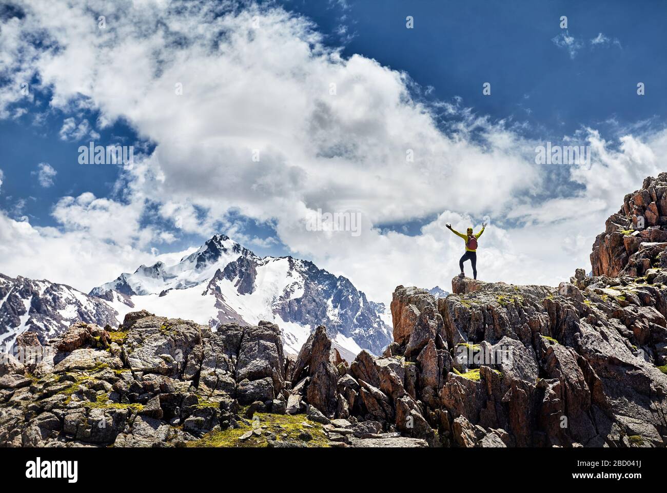 Escursionista in camicia gialla con zaino in piedi sulla roccia con le mani che si alzano godendo la vista delle montagne innevate su sfondo cielo nuvoloso Foto Stock