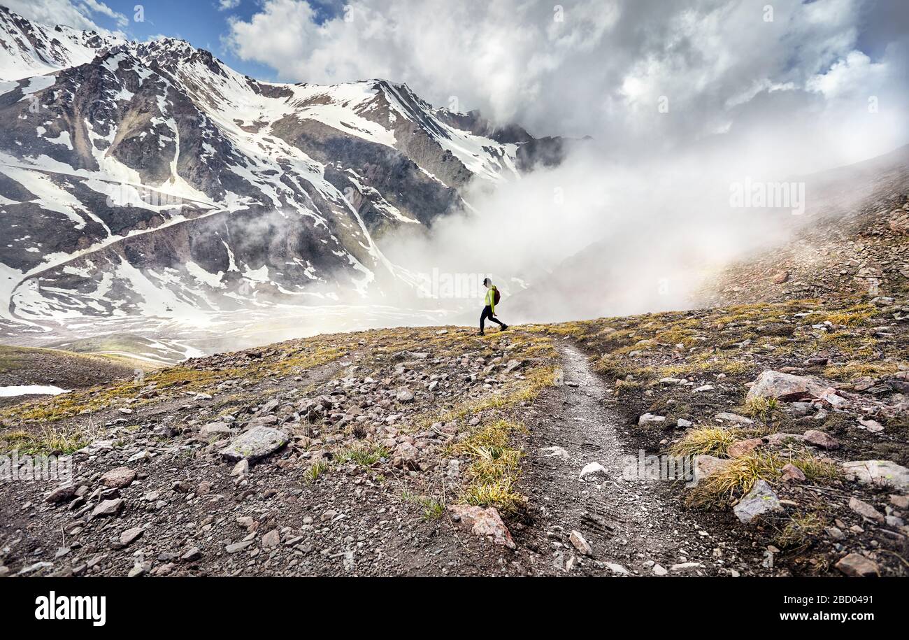 Escursionista in camicia verde con zaino camminando nelle montagne innevate a foggy sfondo cielo Foto Stock