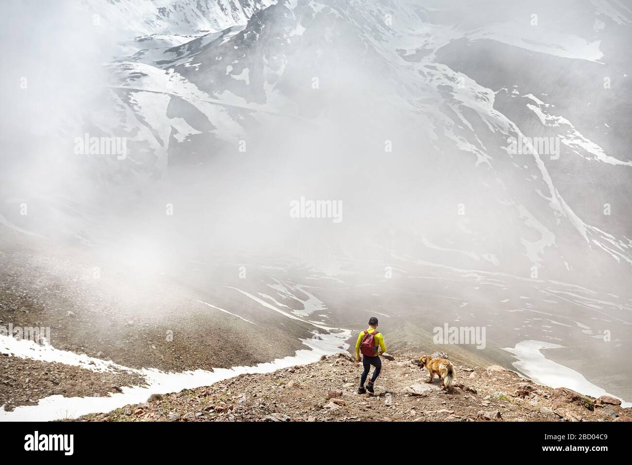 Escursionista in camicia verde con zaino e cane a camminare in montagna innevata a foggy sfondo cielo Foto Stock