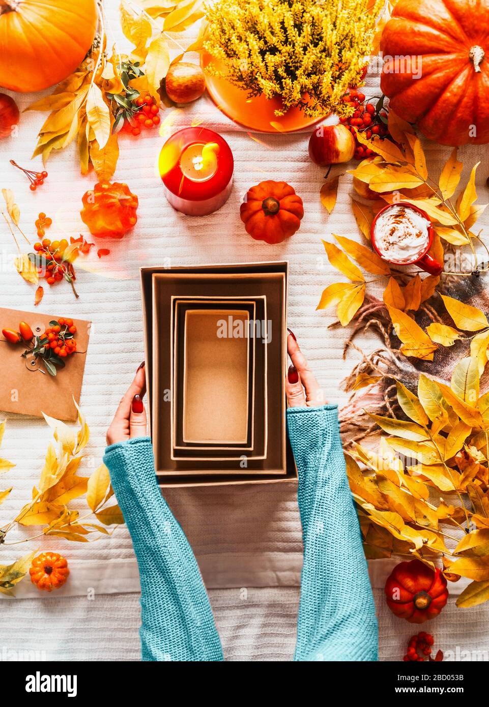 Mani femminili in maglione blu che tiene vuote eco friendly scatole di carta artigianale sulla scrivania con varie zucche, foglie caduta, candele e cappuccino in m rosso Foto Stock