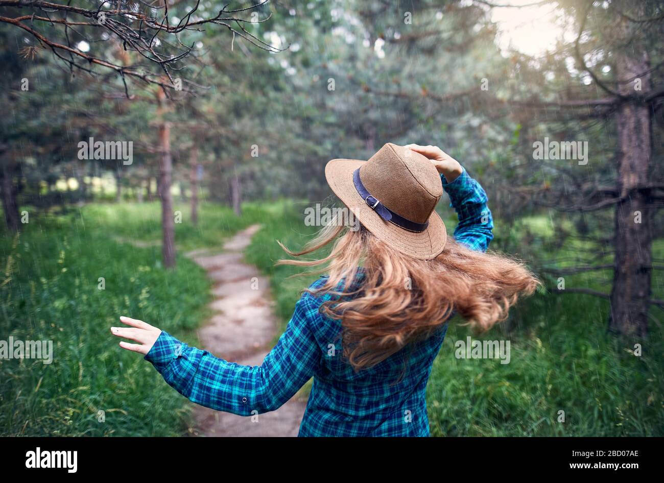 Giovane donna in marrone cappello e verde maglietta controllati con capelli lunghi in esecuzione sotto il nella foresta di alberi di pino Foto Stock