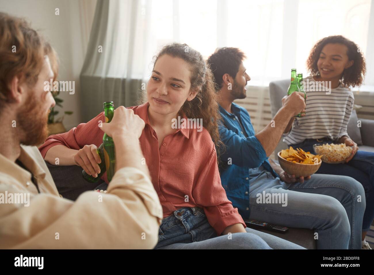 Gruppo multietnico di amici che si aggirano le bottiglie di birra mentre guardano la TV insieme seduti su un comodo divano a casa, concentrarsi sulla giovane coppia a rinunciare Foto Stock