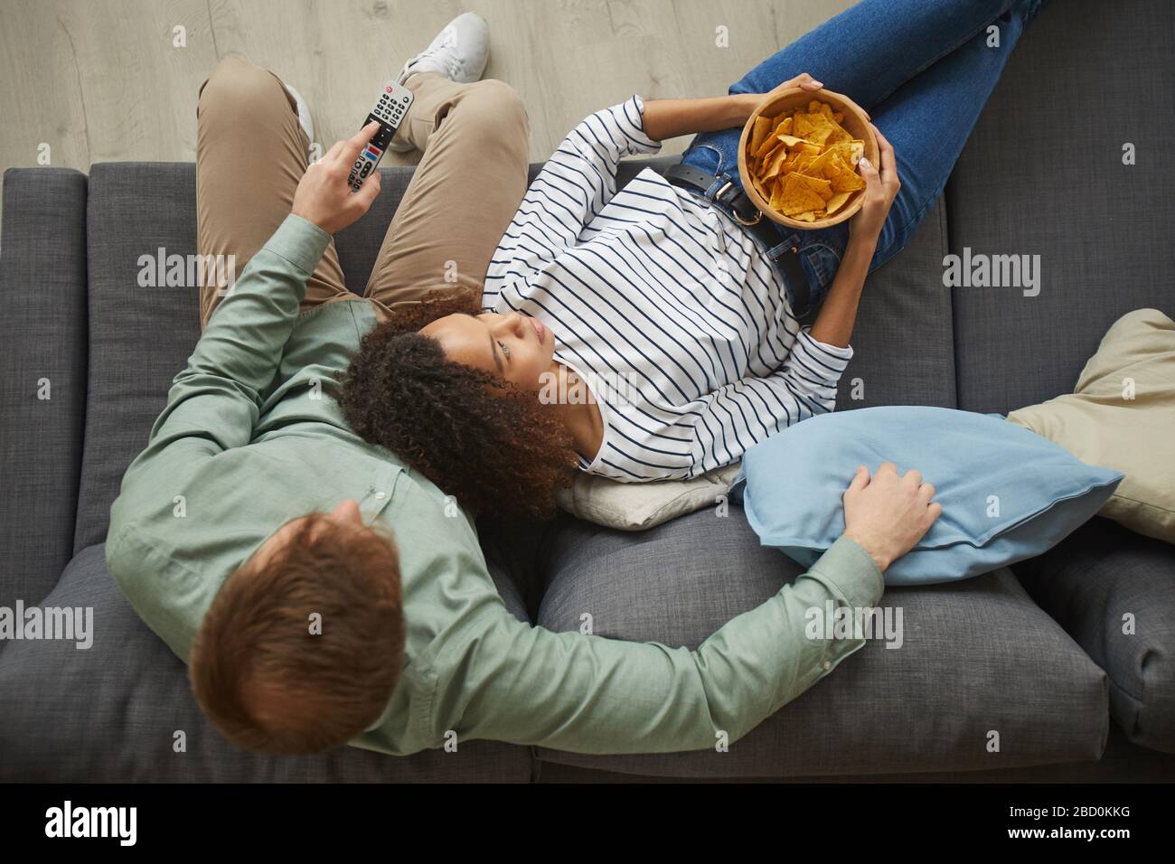 In alto, potrai vedere il ritratto di una giovane coppia di corse miste guardando la TV a casa e mangiare spuntini mentre ti stesi su un comodo divano, uno spazio per copiare Foto Stock