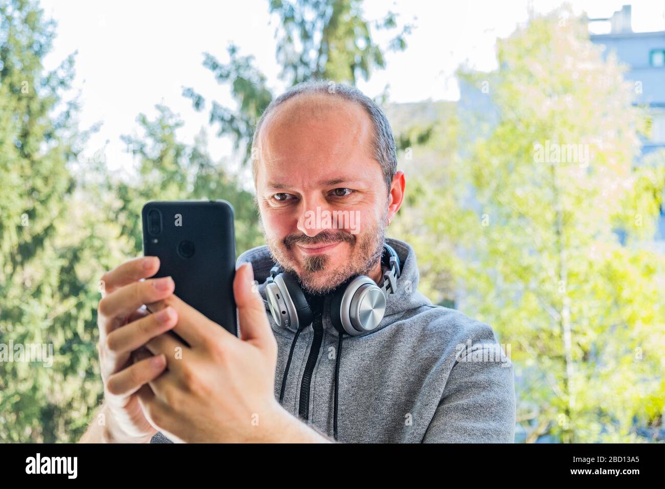 Uomo adulto bearded in felpa con cappuccio con cuffie wireless che prendono il selfie con smartphone nel parco all'aperto, testa e spalle Foto Stock