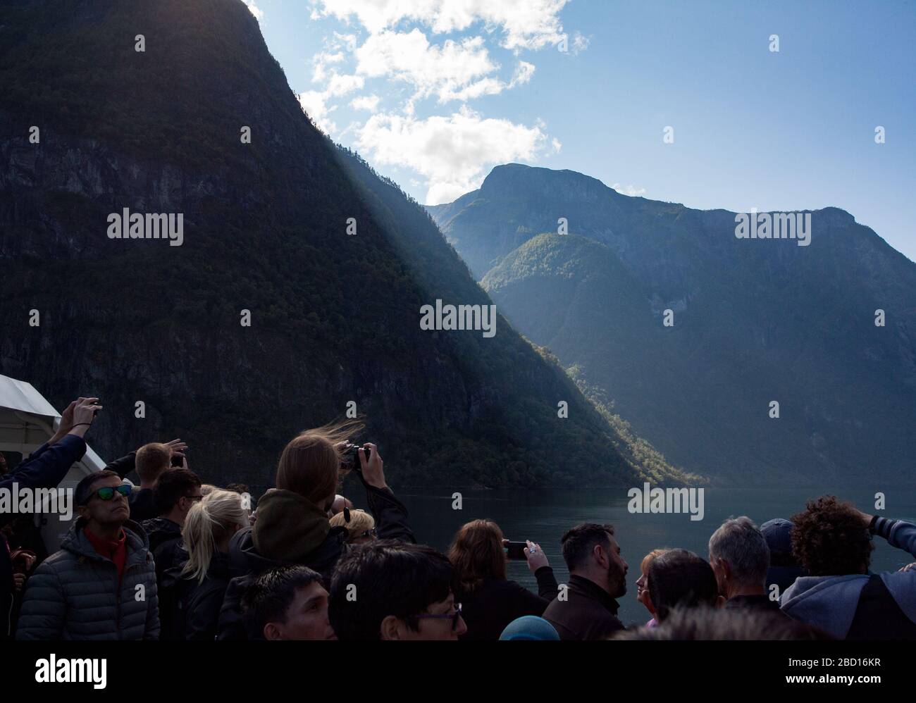 I passeggeri in una crociera sul fiordo sono visibili in primo piano, con la luce del sole che si affaccia su una scogliera coperta da alberi e si affaccia su un fiordo Foto Stock
