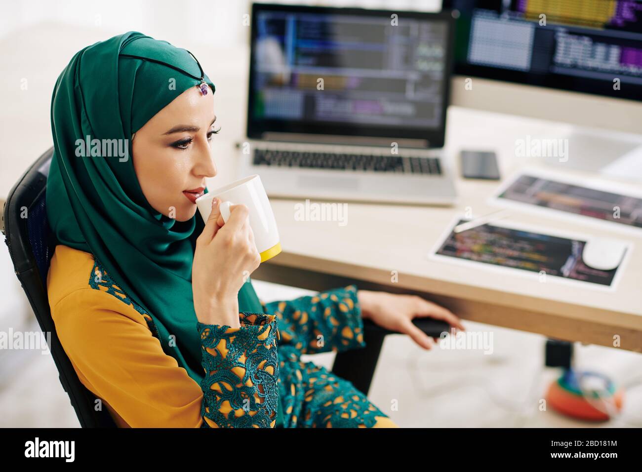 Pensiva giovane donna musulmana seduta alla scrivania e bevendo una tazza grande di tè o caffè Foto Stock