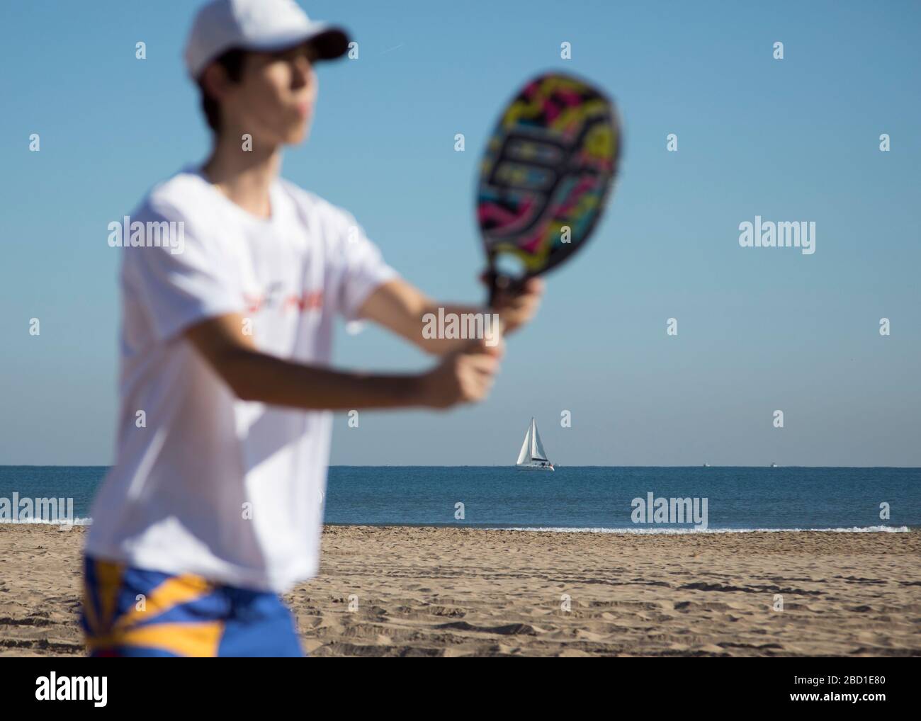 Una barca a vela vista in lontananza come l'uomo gioca Beach Tennis con un cielo blu soleggiato. Foto Stock