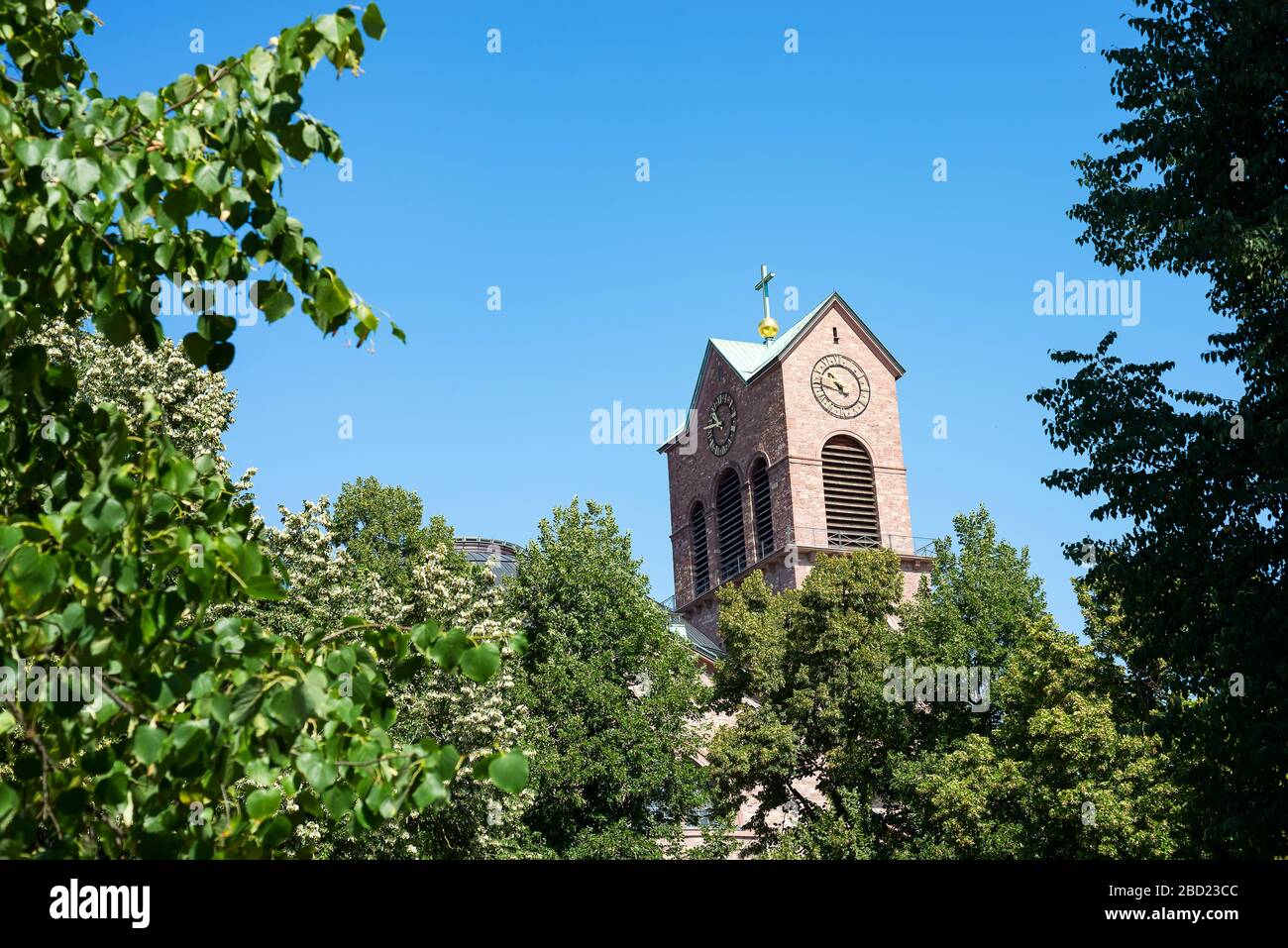 Karlsruhe, Germania - 27 luglio 2018. Vecchio orologio sul campanile della chiesa di Santo Stefano tra alberi verdi cime su uno sfondo di cielo azzurro in città Foto Stock