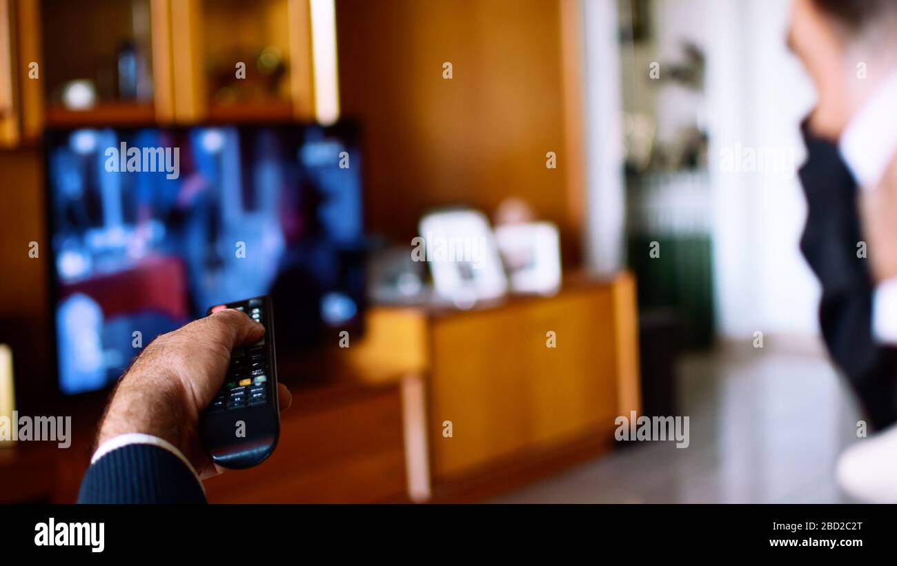 Uomo che indossa la maschera protettiva durante la quarantena covid-19 utilizzando il telecomando per cambiare il programma TV. Isolamento a casa. Foto Stock