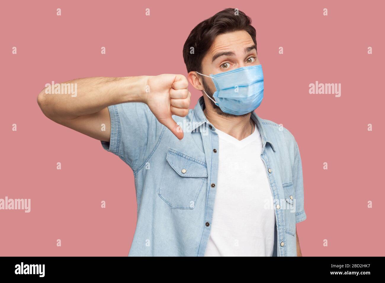 No, non mi piace. Ritratto di giovane insoddisfatto con maschera medica chirurgica in camicia blu in piedi, pollici giù e guardando la macchina fotografica . interni studio s. Foto Stock