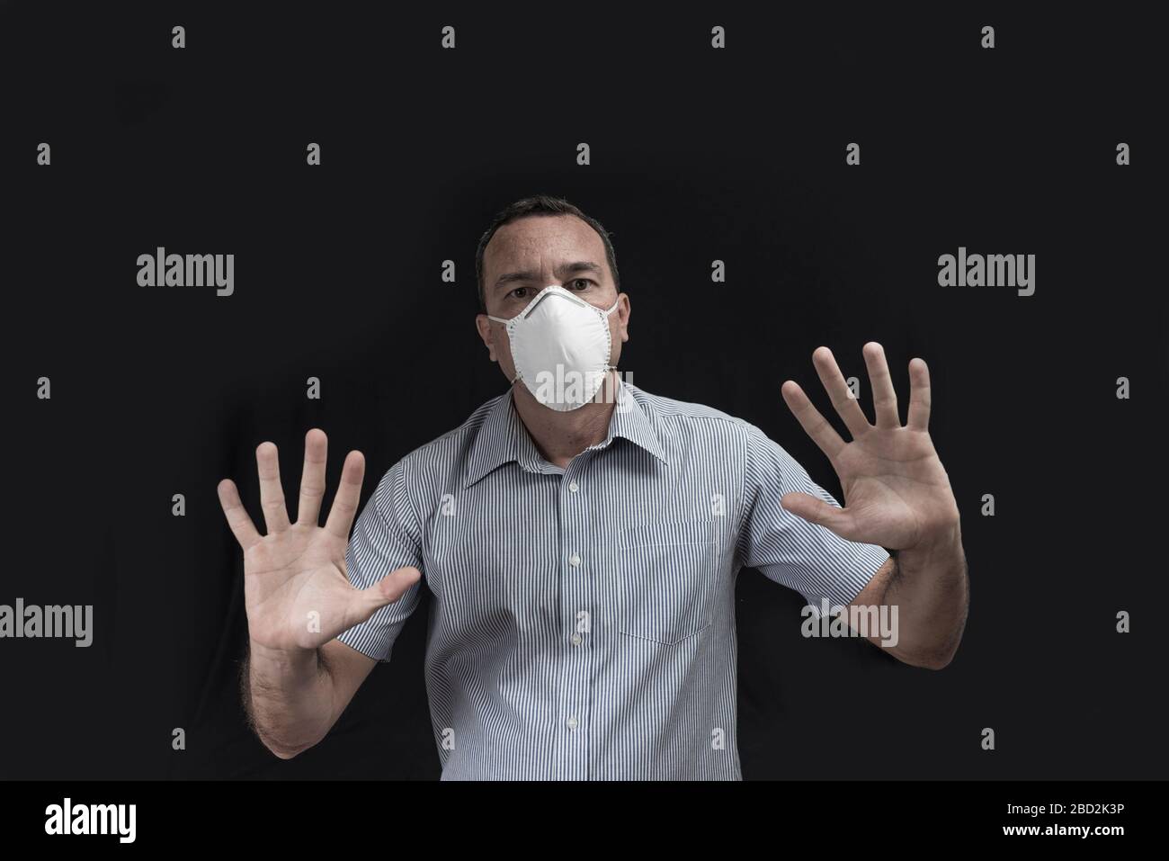 Un uomo bianco che indossa una maschera protettiva che rende un gesto di stare lontano con entrambe le mani su uno sfondo nero Foto Stock