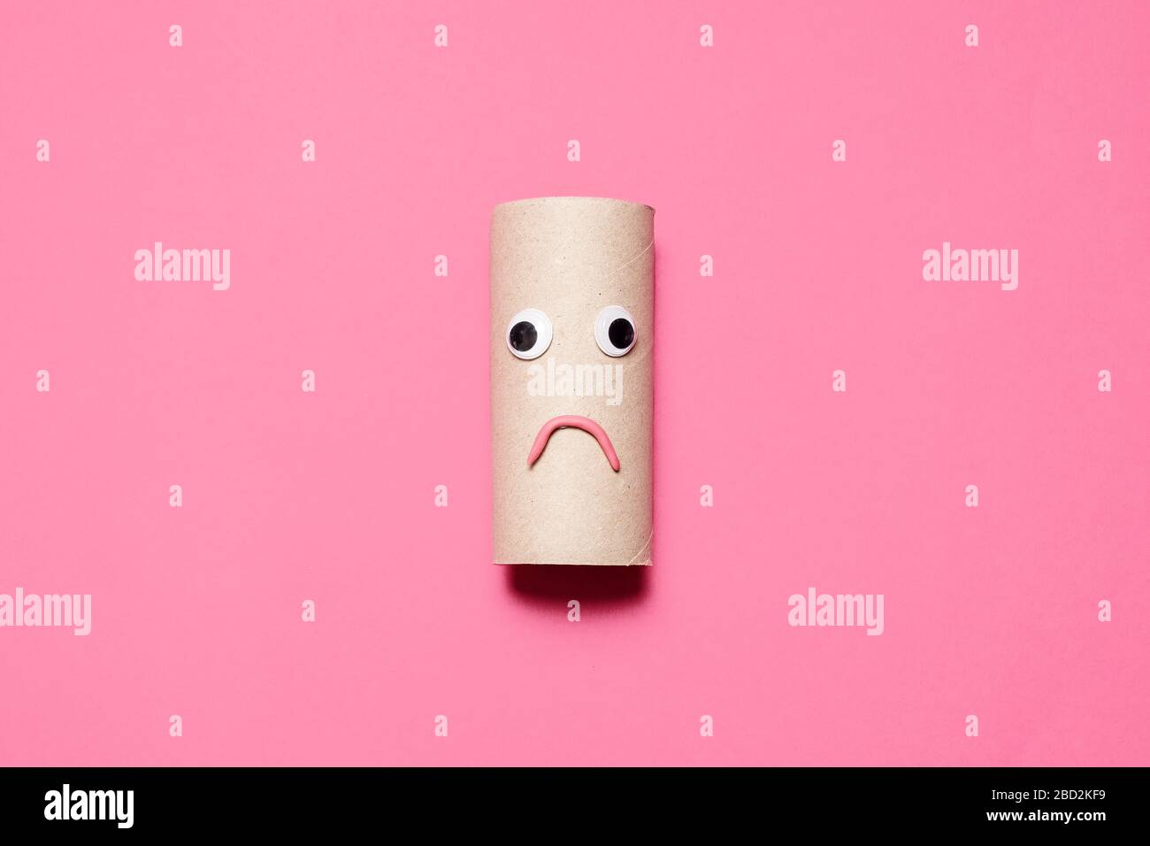 Rotolo vuoto triste e accigliato di carta igienica con occhi e bocca googly su uno sfondo rosa con spazio per le copie e spazio per il testo. Foto Stock