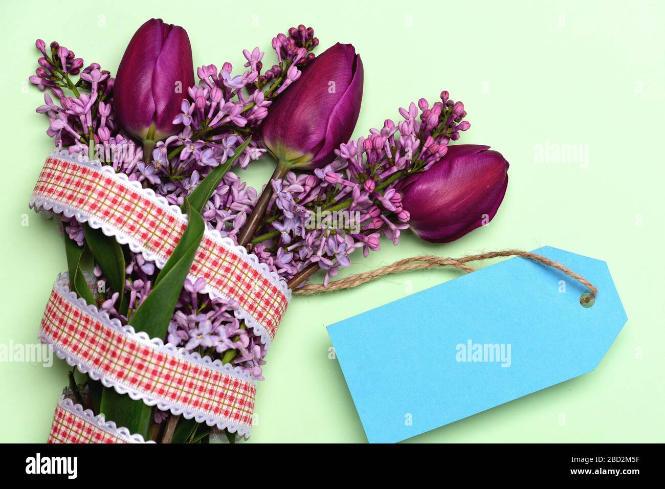Mazzo di lilla e tulipani con nastro rustico in rosso e bianco con etichetta blu brillante isolata su sfondo chiaro. Vista dall'alto e spazio copia, concetto di regalo e vacanza primaverile Foto Stock