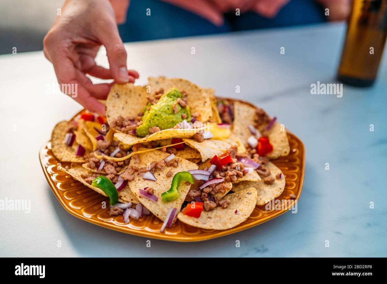 Persone mani immersione giallo nacho patatine guarnite con manzo macinato, guacamole, formaggio fuso, peperoni e foglie di coriandolo in piastra su pietra bianca Foto Stock