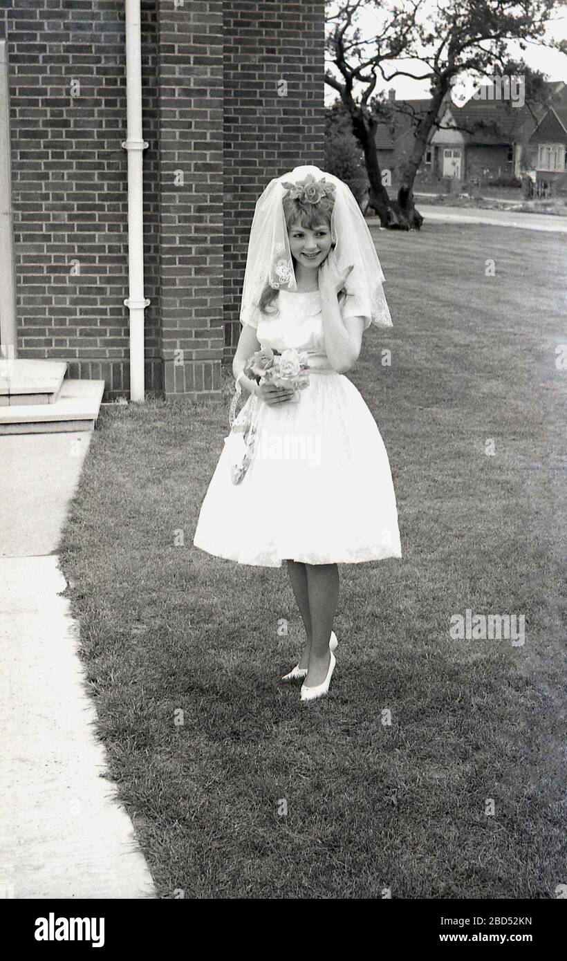 Anni '60, storico, sposa adolescente nel suo abito da sposa in piedi fuori della chiesa moderna per una fotografia, Inghilterra, Regno Unito. In quest'epoca come in quelle precedenti, le persone si sposarono con un'età molto più giovane delle generazioni successive. Foto Stock