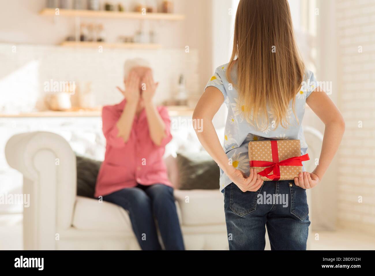 Bambina che nasconde il presente per la sua nonna dietro la schiena a casa, spazio vuoto Foto Stock