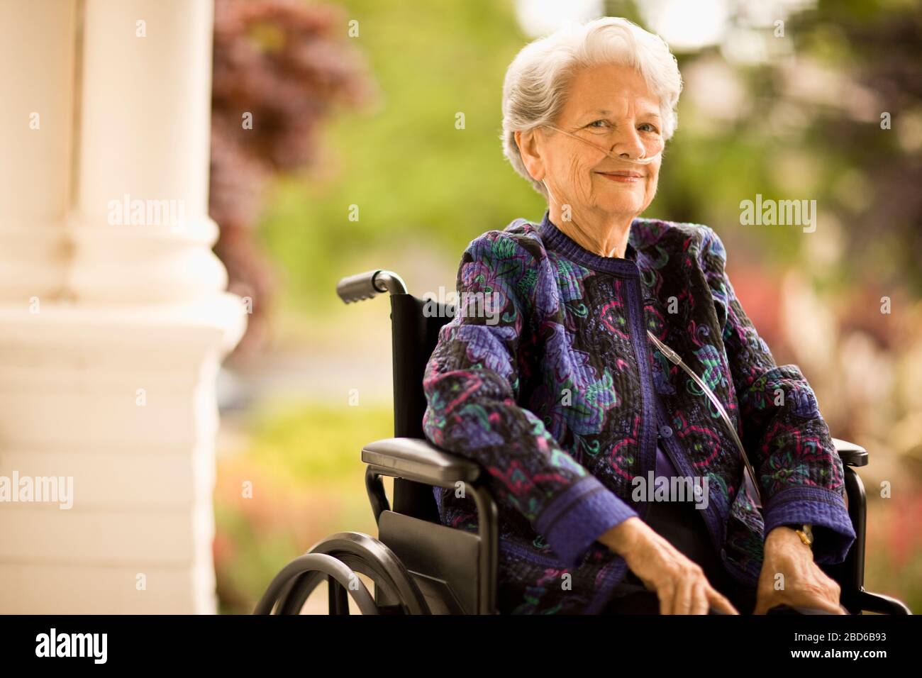 Donna anziana con tubo per ossigeno (cannula) siede su una sedia a rotelle su un portico e si posa per un ritratto. Foto Stock