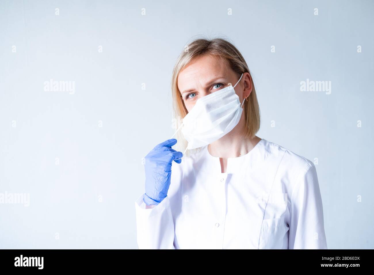 Medico chirurgo femmina bionda in uniforme bianca, guanti protettivi blu indossare su maschera medica su grigio. Assistenza sanitaria, soggiorno a casa. copyspace Foto Stock