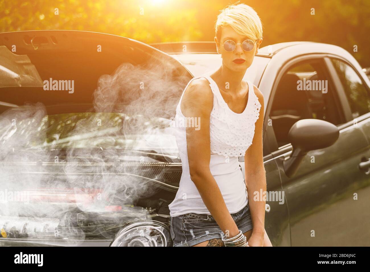 Giovane donna triste in piedi vicino alla strada, vicino alla macchina rotta nel mezzo del nulla. Fumo proveniente dal surriscaldamento del motore. Servizio di traino in attesa di auto. Foto Stock