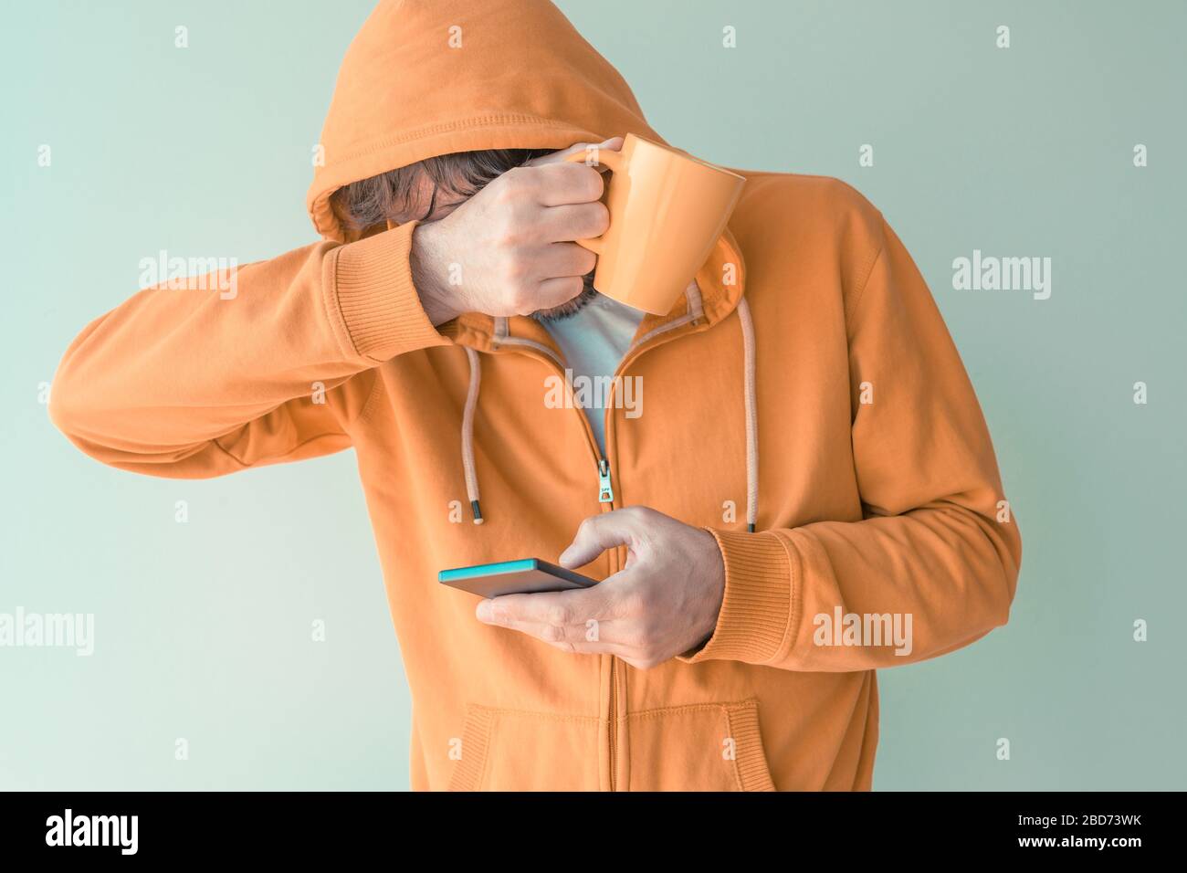 Uomo che beve il caffè e che usa il telefono cellulare al mattino, adulto maschio con cappuccio giallo che tiene tazza di caffè e smartphone, messa a fuoco selettiva Foto Stock