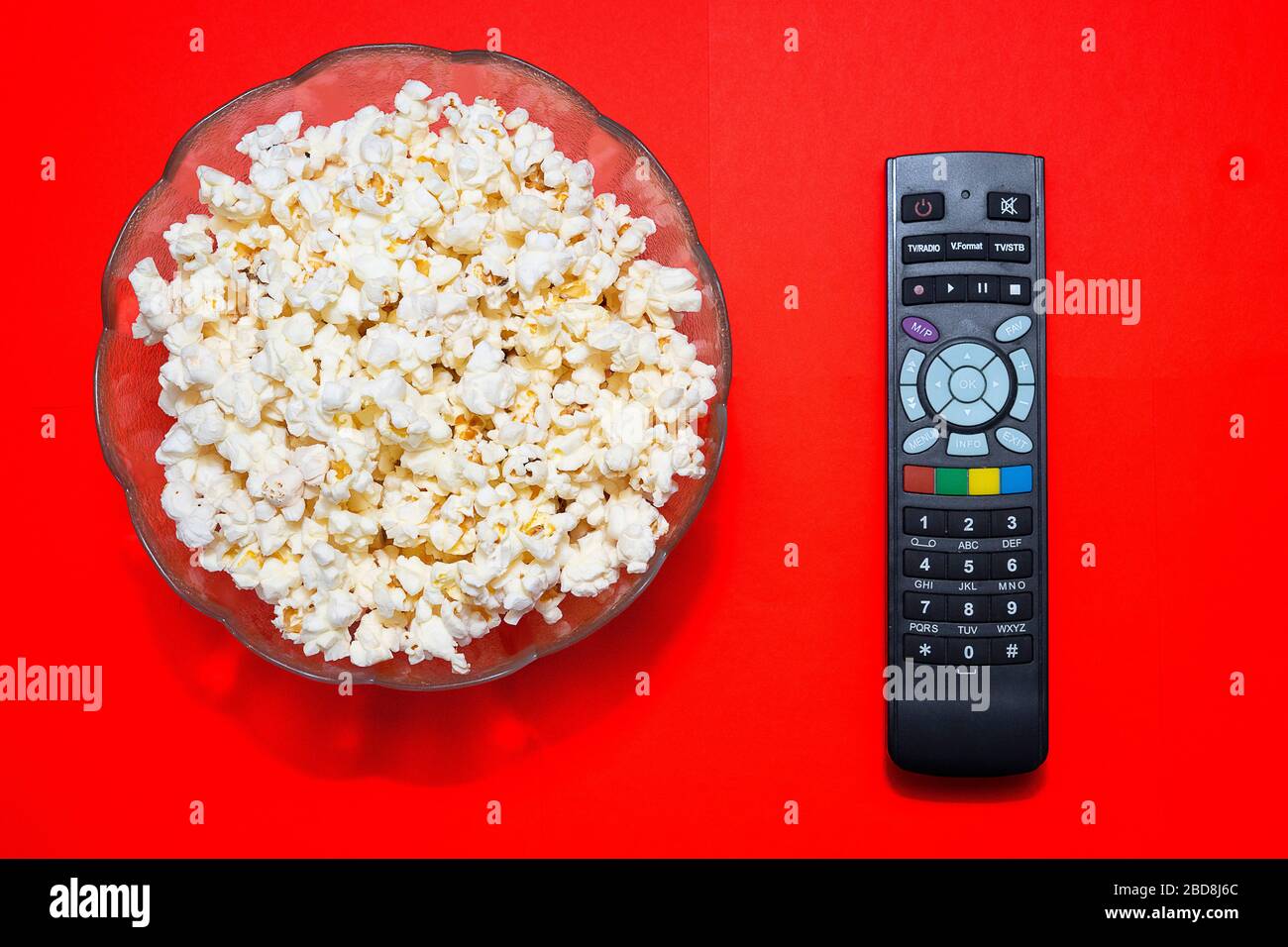 Ciotola con popcorn salato accanto a un telecomando tv su sfondo rosso. Concetto di controllo popcorn e tv. Foto Stock