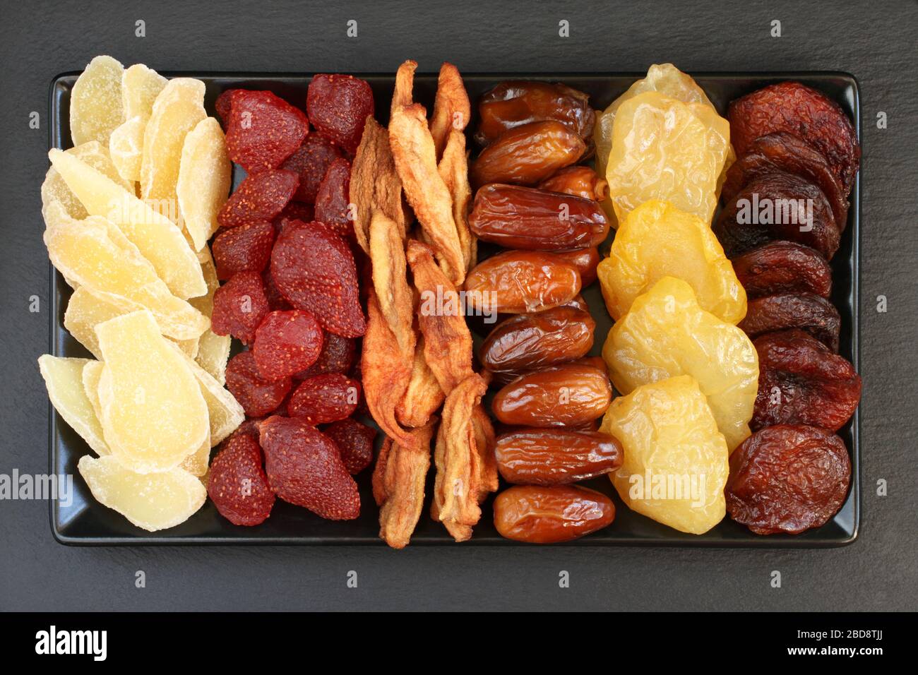 Сandied fette di zenzero e fragole, fette di pera essiccate, datteri, pesche e albicocche giacciono su un piatto di ceramica nera su una superficie di pietra nera Foto Stock