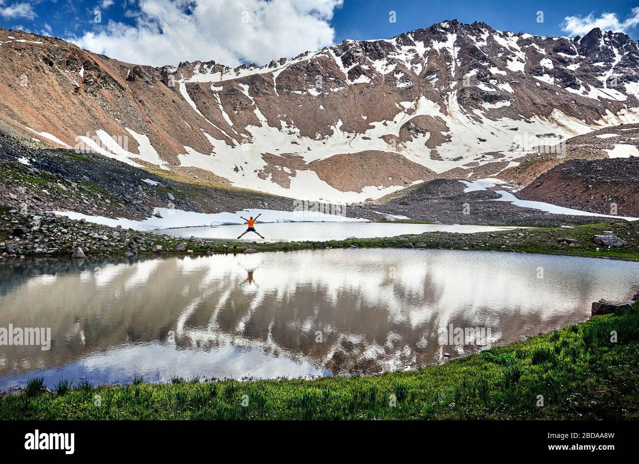 Escursionista in maglietta arancione jumping con origine mano vicino lago nelle montagne innevate Foto Stock