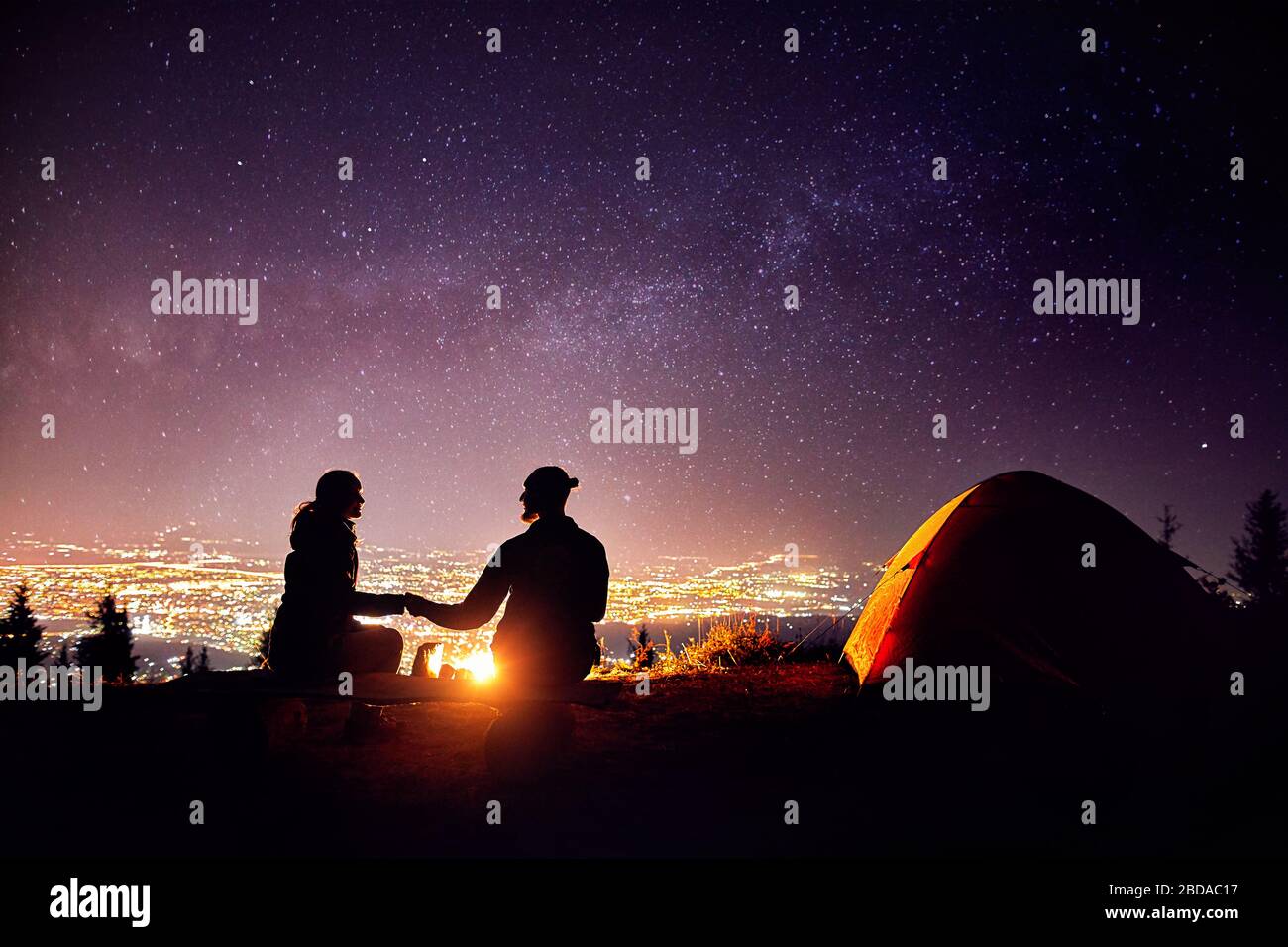 Coppia felice in silhouette seduto vicino al fuoco e tenda arancione. Cielo notturno con via lattea stelle e luci della città a sfondo. Foto Stock