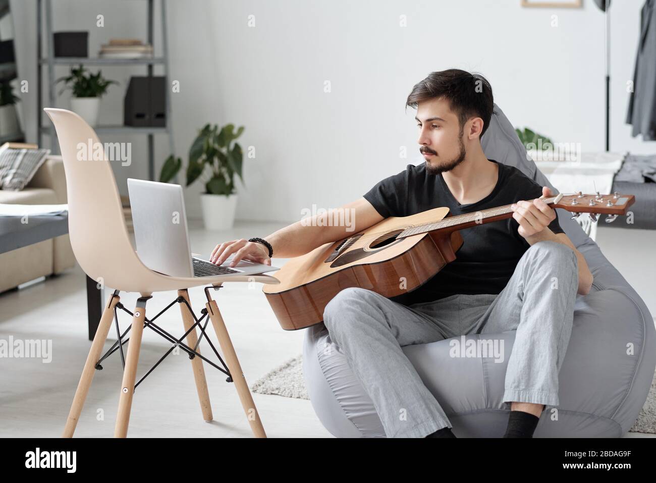 Giovane uomo serio seduto in una borsa di fagioli e utilizzando il laptop mentre imparano a suonare la chitarra a casa durante la quarantena Foto Stock