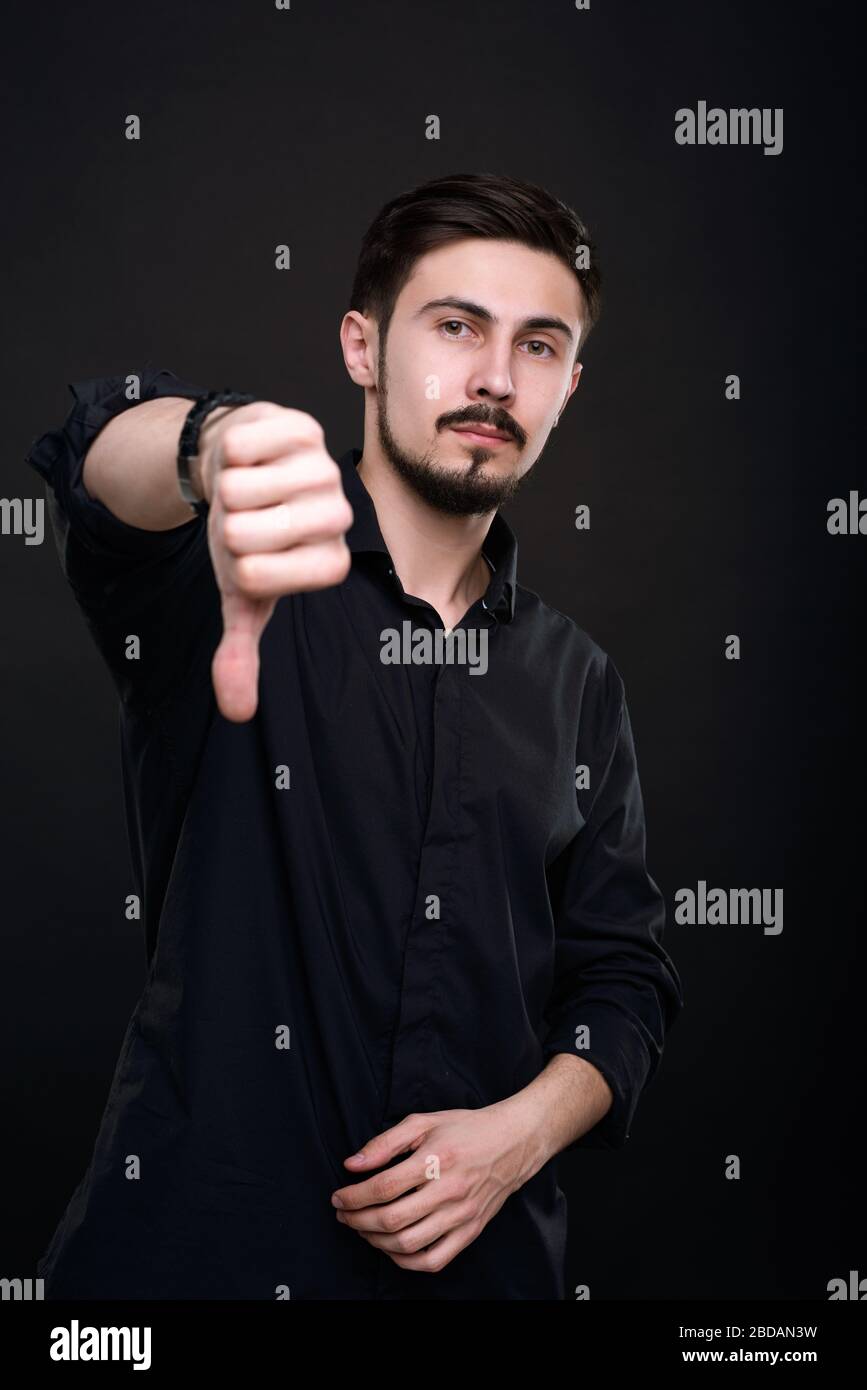 Ritratto di serious bel giovane uomo in camicia nera che mostra il pollice giù mentre dà il grado difettoso contro lo sfondo scuro isolato Foto Stock