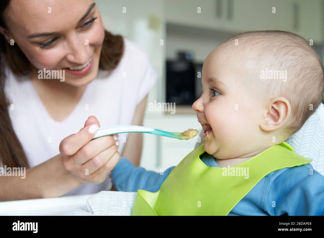 Sorridente 8 mese vecchio Baby Boy a casa in alta sedia essendo alimentato cibo solido da madre con cucchiaino Foto Stock