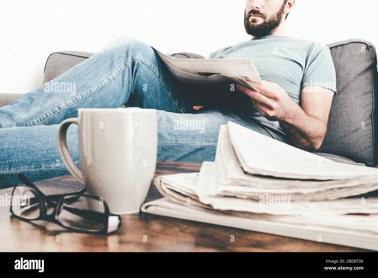 uomo che si rilassa sul divano leggendo un giornale con una pila di giornali e una tazza di caffè in primo piano Foto Stock