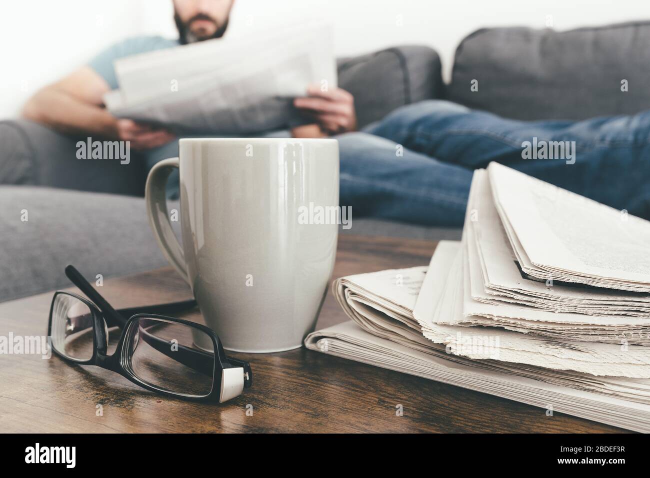 uomo che si rilassa sul divano leggendo un giornale con una pila di giornali e una tazza di caffè in primo piano Foto Stock