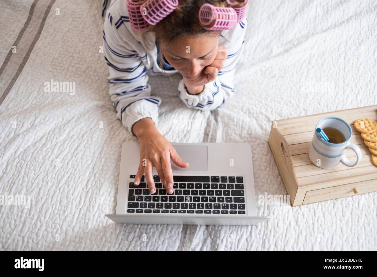 Donna con arricciacapelli sdraiata sul letto e che utilizza un computer portatile Foto Stock