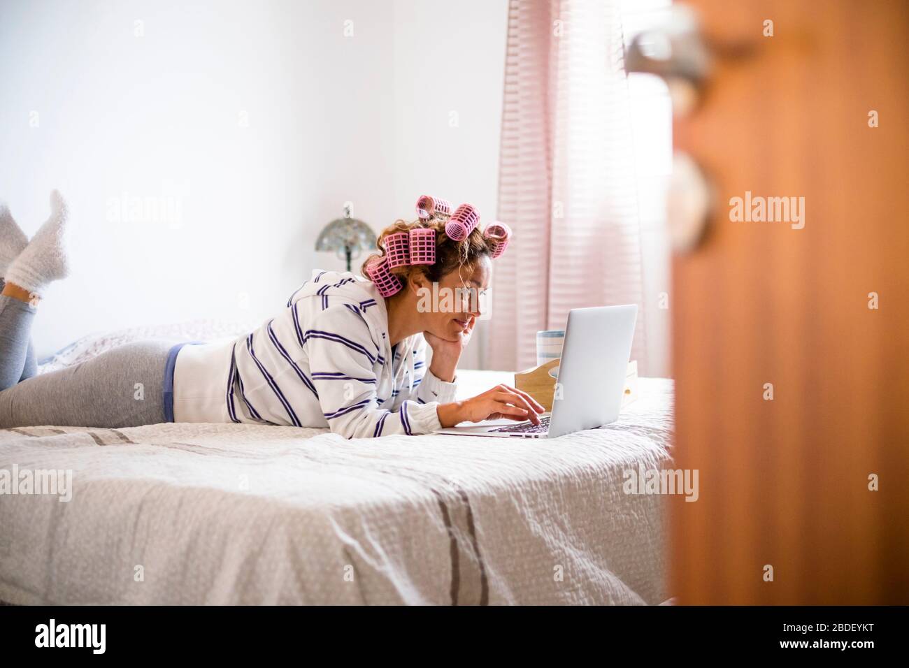 Donna con arricciacapelli sdraiata sul letto e che utilizza un computer portatile Foto Stock