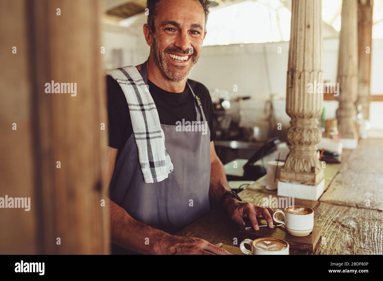Uomo medio adulto in grembiule guardando la macchina fotografica e sorridendo mentre si sta dietro il bancone. Sorridente barista maschile in una caffetteria. Foto Stock