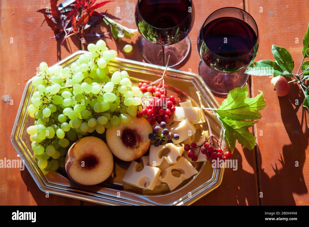 Due bicchieri di vino rosso e formaggio a tavola, uve fresche, pesche e frutti di bosco su tavola. Posizione esterna Foto Stock