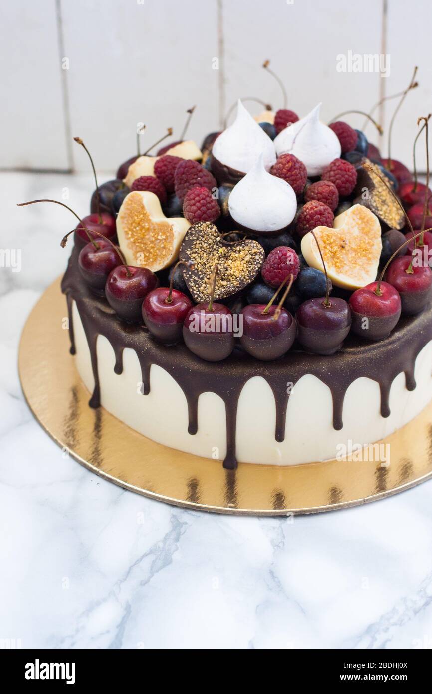 Cheesecake alla vaniglia decorata con cioccolato fondente fuso, frutti di bosco freschi, ciliegie, cuori di cioccolato e meringhe. Sfondo rustico bianco. Foto Stock