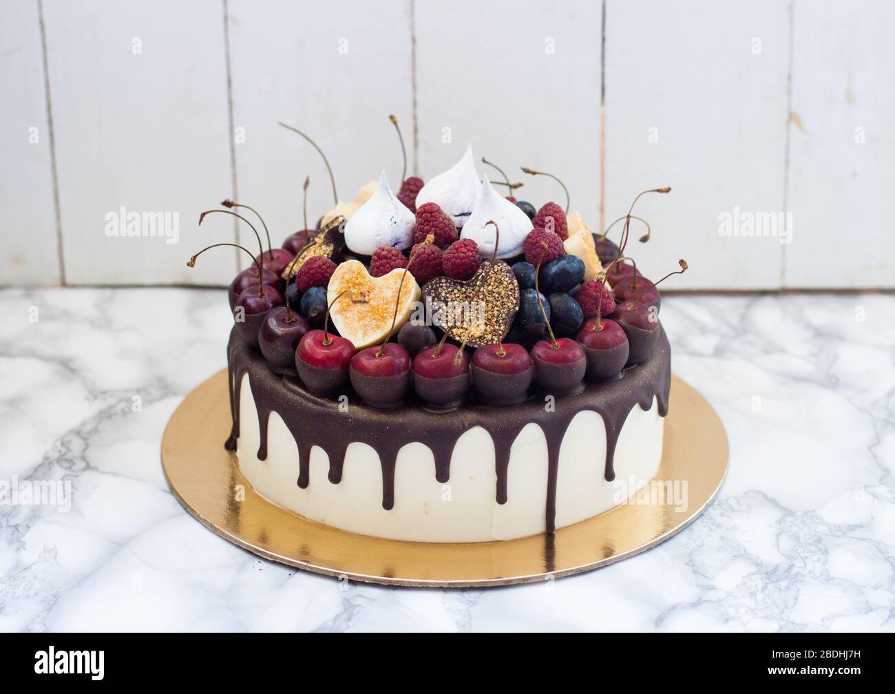 Cheesecake alla vaniglia decorata con cioccolato fondente fuso, frutti di bosco freschi, ciliegie, cuori di cioccolato e meringhe. Sfondo rustico bianco. Foto Stock