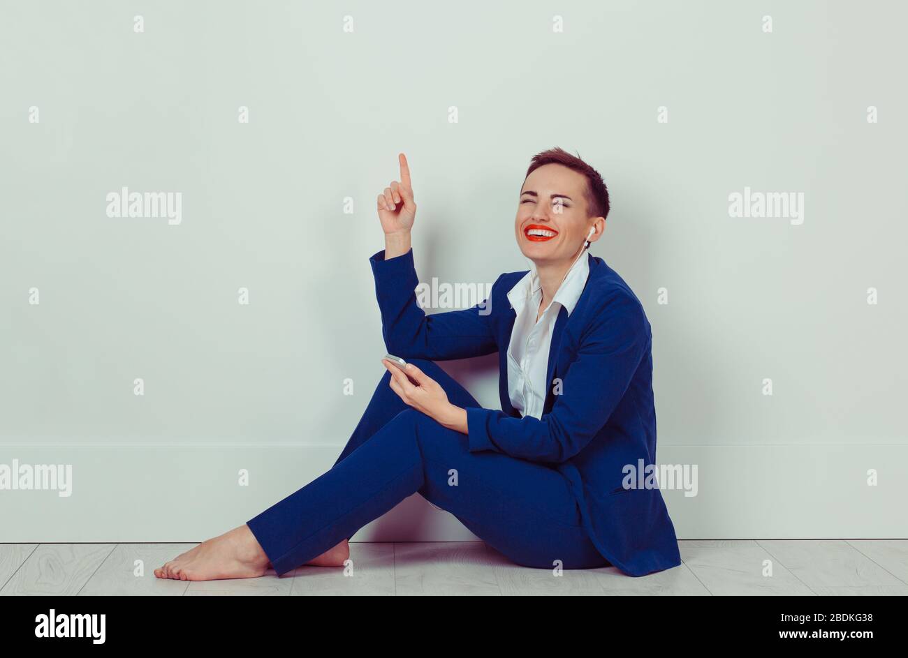 Donna sorridente ascoltare musica in cuffie sul pavimento nella sua stanza, studio isolato bianco muro di mattoni. Foto Stock