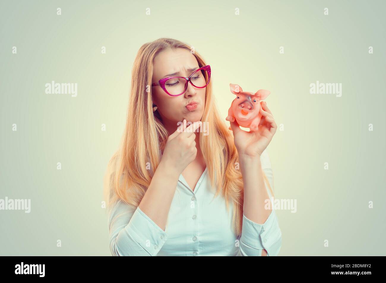 Il ritratto di closeup ha sottolineato la giovane donna che guarda il pensiero della banca piggy, che decide meditando i piani finanziari isolato sfondo verde. Difficoltà di denaro Foto Stock