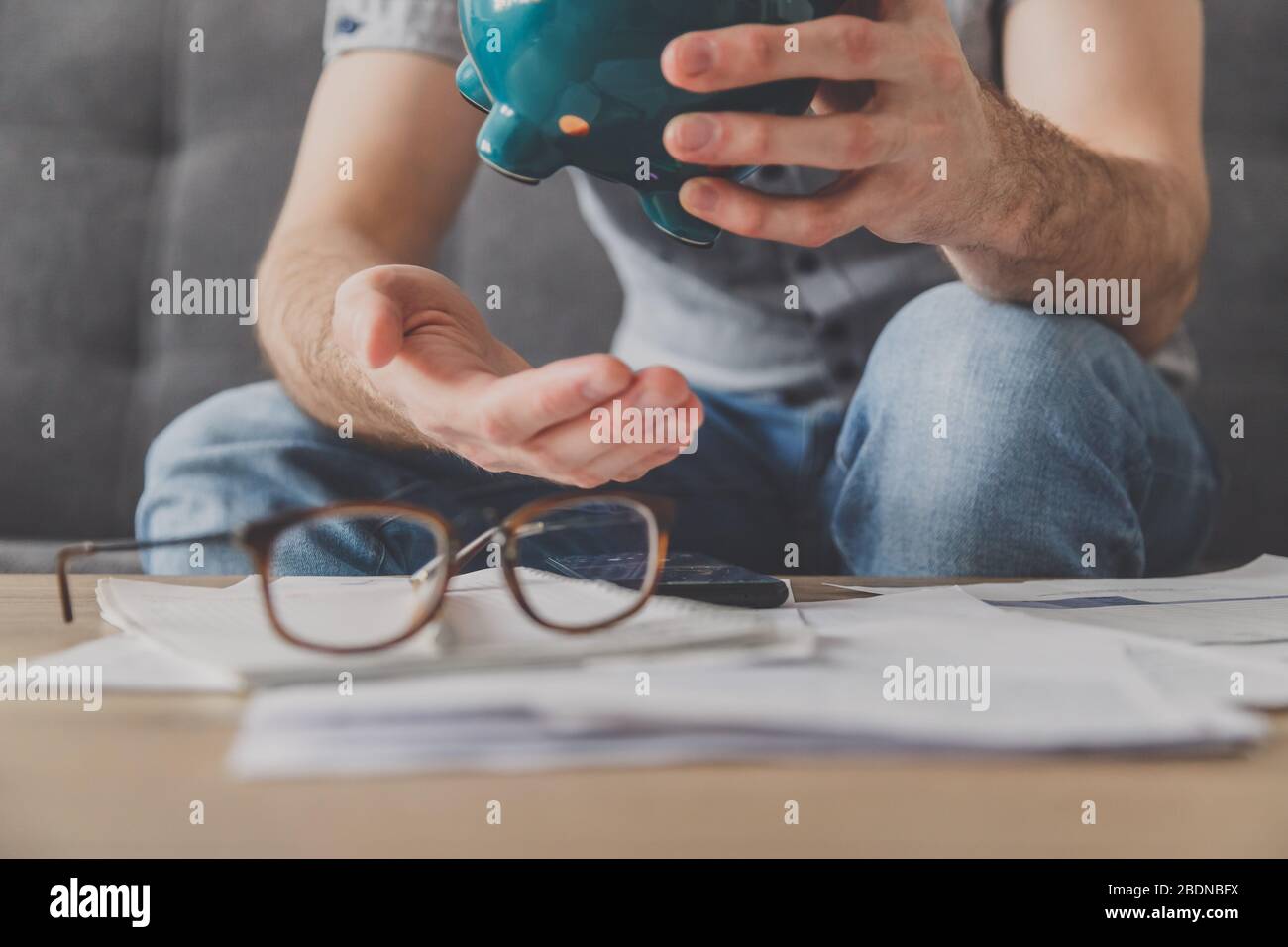 L'uomo seduto ad un tavolo pieno di fatture non pagate scrolla l'ultimo centesimo dalla banca piggy. Spendi i tuoi ultimi risparmi. Disoccupazione, povertà, fallimento Foto Stock