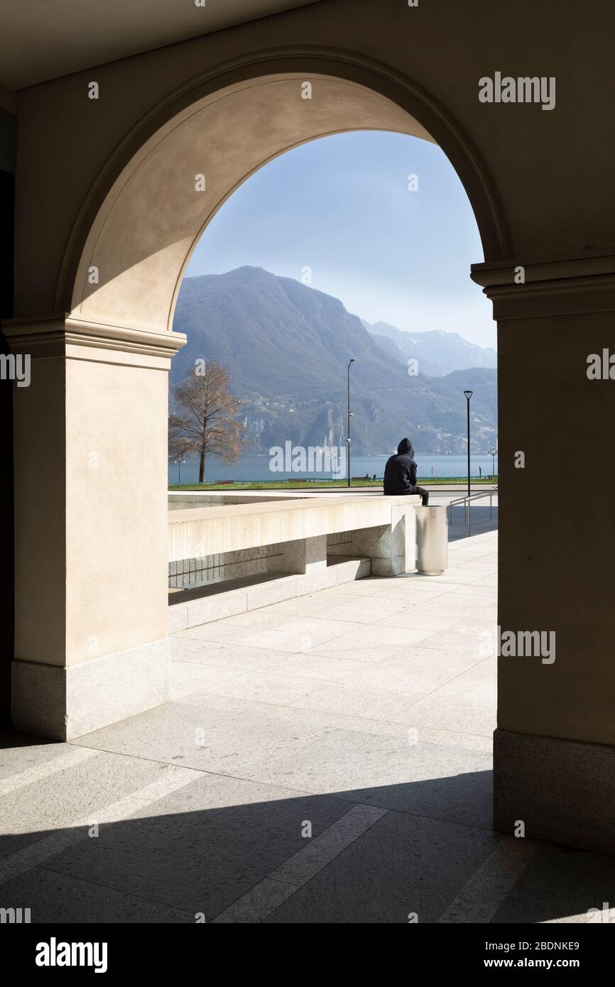 Un uomo è seduto sul muro e guarda il lago dalla città di Lugano. Un arco fa la cornice. Giorno di sole. Foto Stock