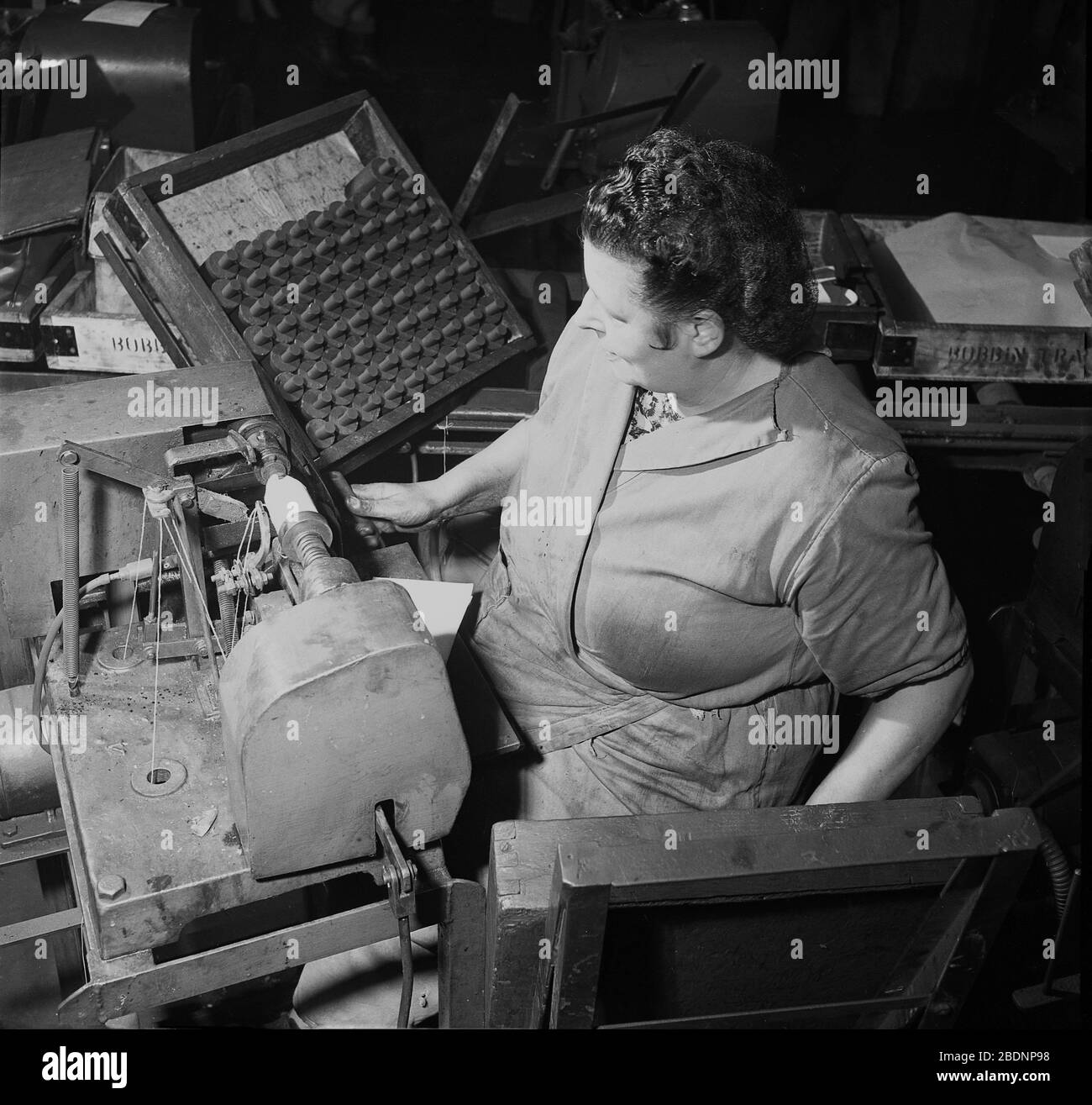 Anni '50, storico, una lavoratrice femminile al negozio che indossa tute sedute a macchinari e accanto a un vassoio di bobine in legno in una fabbrica industriale che produce batterie zinco-carbonio, Londra, Inghilterra. Nel Regno Unito del dopoguerra, molte delle donne che sono andate nelle fabbriche per aiutare lo sforzo di guerra sono rimaste in loro dopo la guerra, mentre hanno guadagnato soldi per sostenere le loro famiglie. Foto Stock