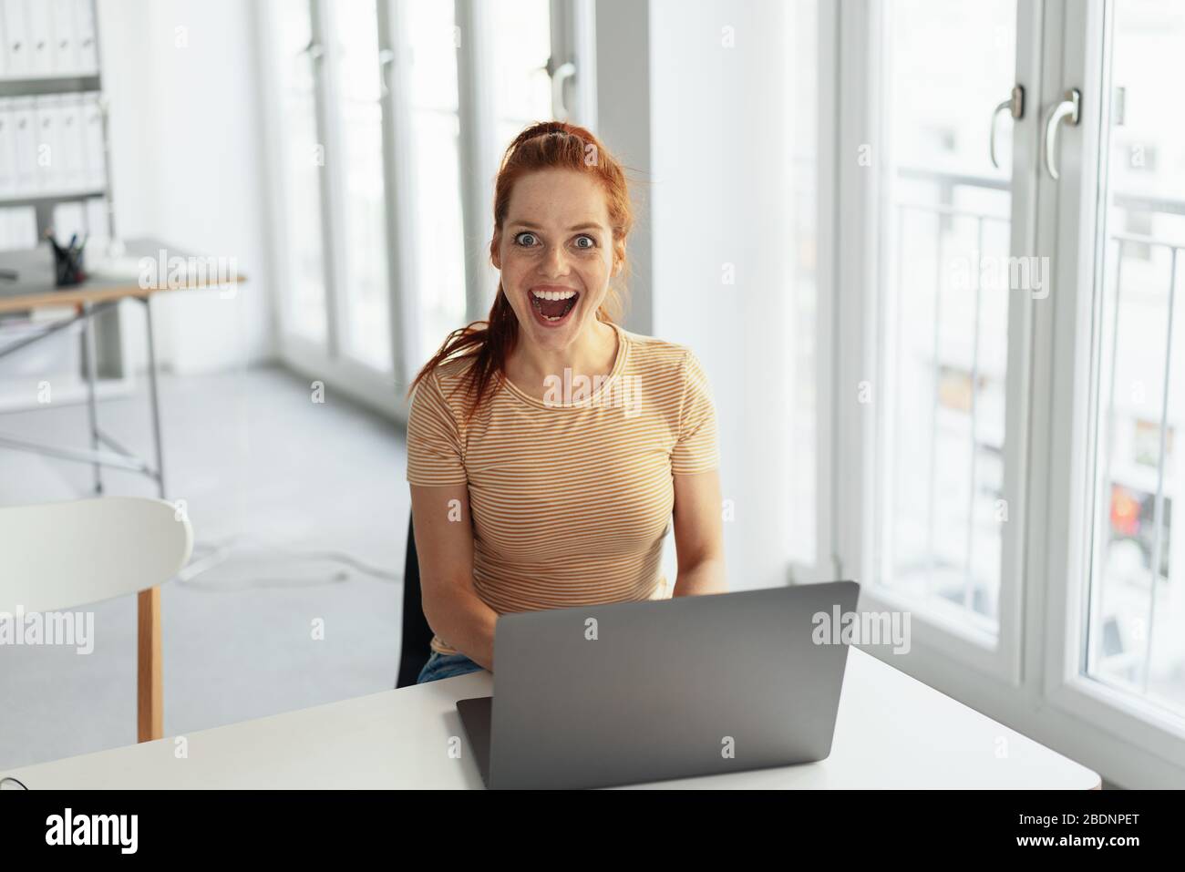 Sorpresi giovane donna d'affari sorridente alla macchina fotografica con gli occhi larghi eccitati e la bocca aperta mentre siede al suo laptop in un ufficio chiave alta con spazio di copia Foto Stock