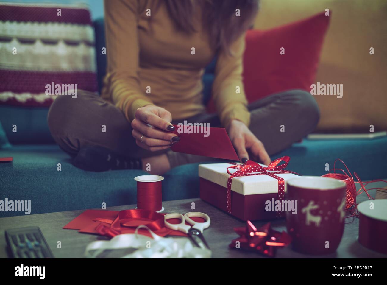 Donna che prepara regali di Natale a casa: È seduta sul divano e l'aggiunta di una cartolina di Natale ad un regalo Foto Stock
